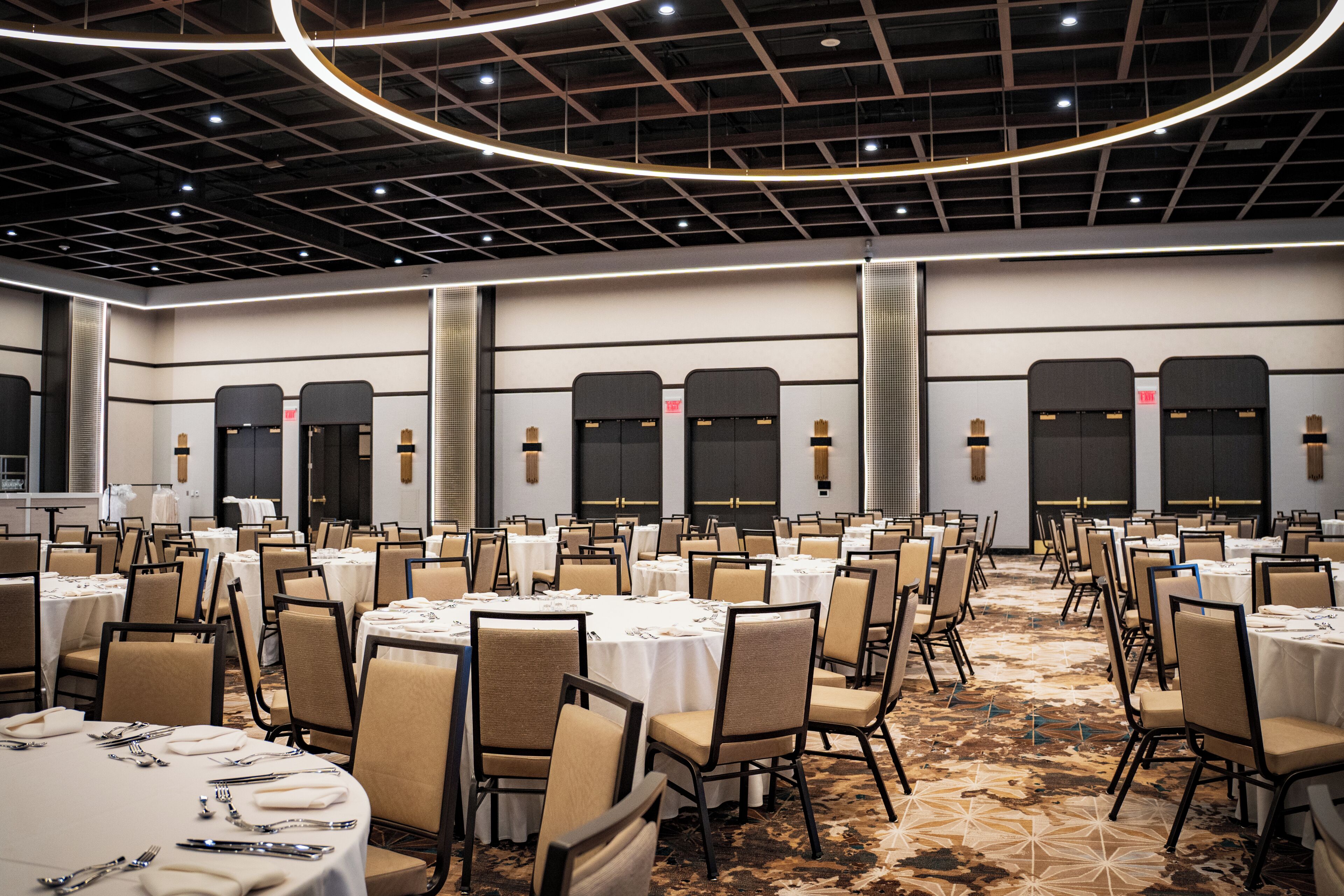 A large banquet hall with round tables and chairs, a patterned carpet, and a high ceiling with a grid design.
