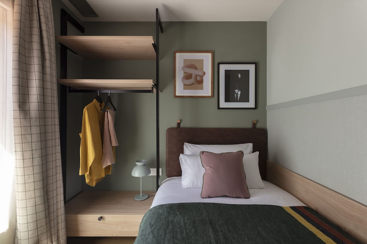 A neatly arranged hotel room with a bed, a bedside table, a hanging coat rack, and a wall-mounted shelf.