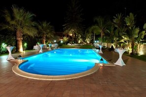 A nighttime swimming pool area with a curved pool, surrounded by palm trees and white tables with umbrellas. - Villa Luca (Sant'Agata di Militello)