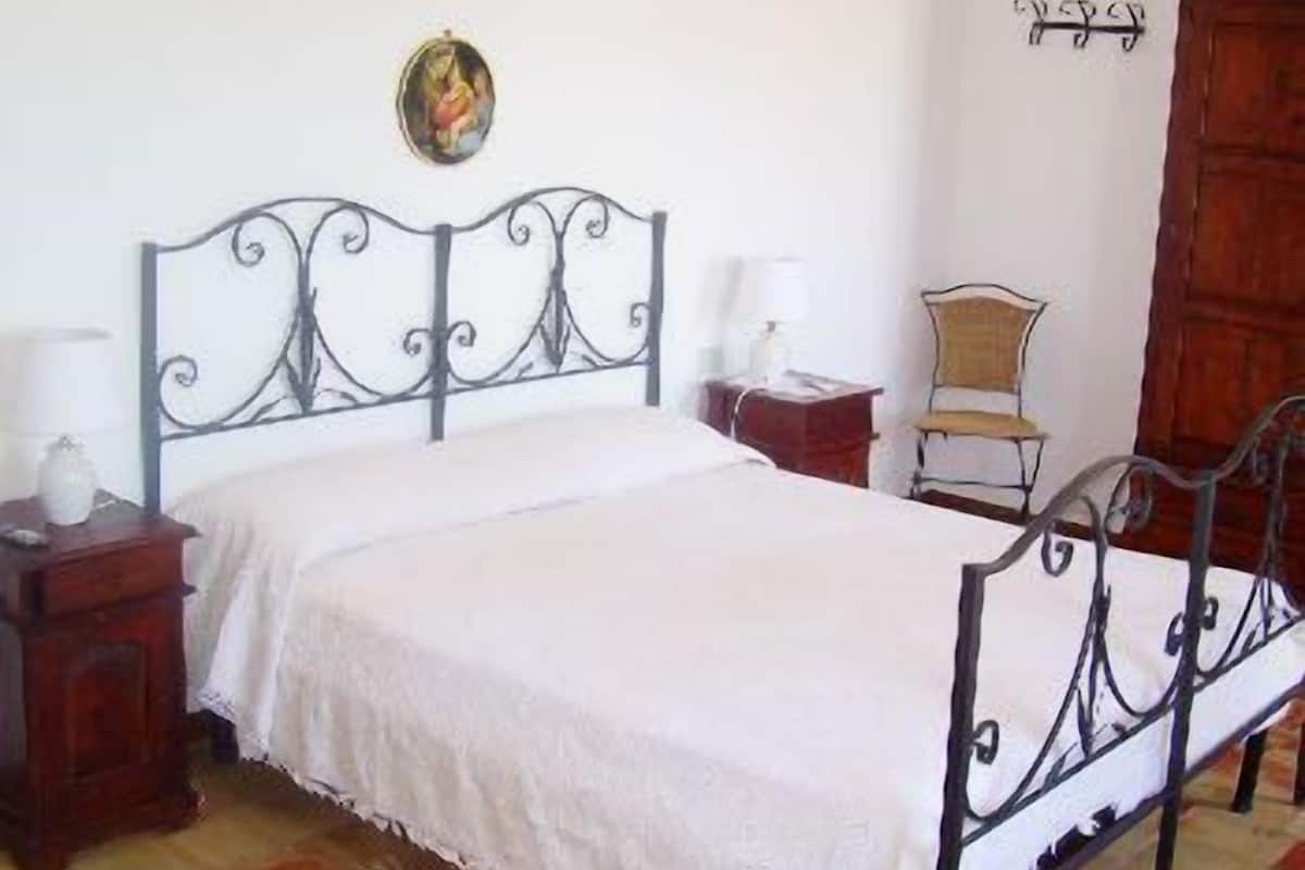 A bedroom with a metal bed frame, a wooden nightstand, a chair, and a wooden wardrobe.