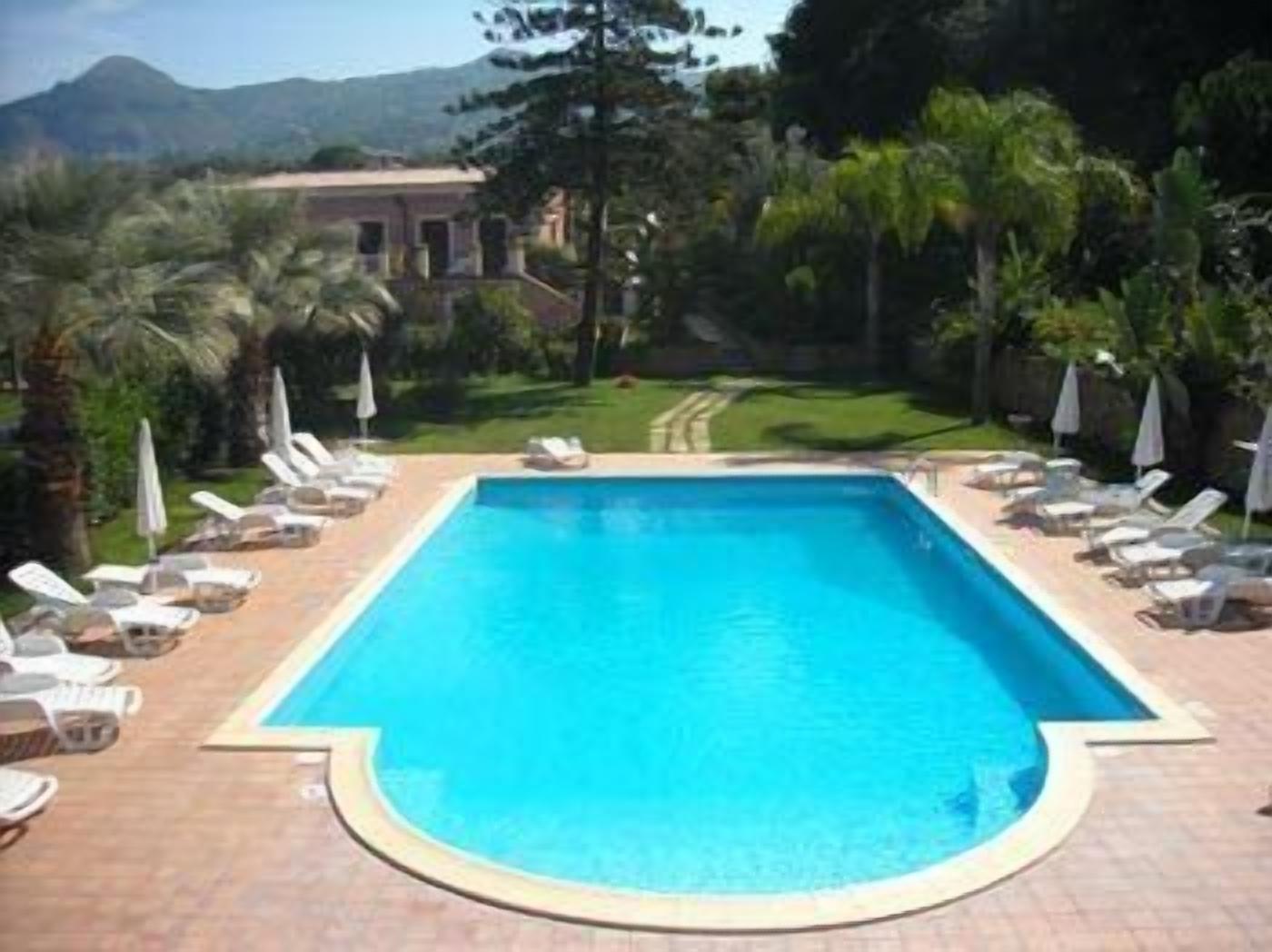A rectangular swimming pool with a tiled deck, surrounded by lounge chairs and umbrellas, set against a backdrop of greenery and distant mountains.