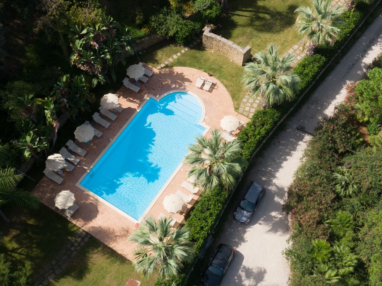 Eine Vogelperspektive zeigt einen Swimmingpool, umgeben von Liegestühlen und Palmen, mit einem gepflasterten Weg und einem parkenden Auto.