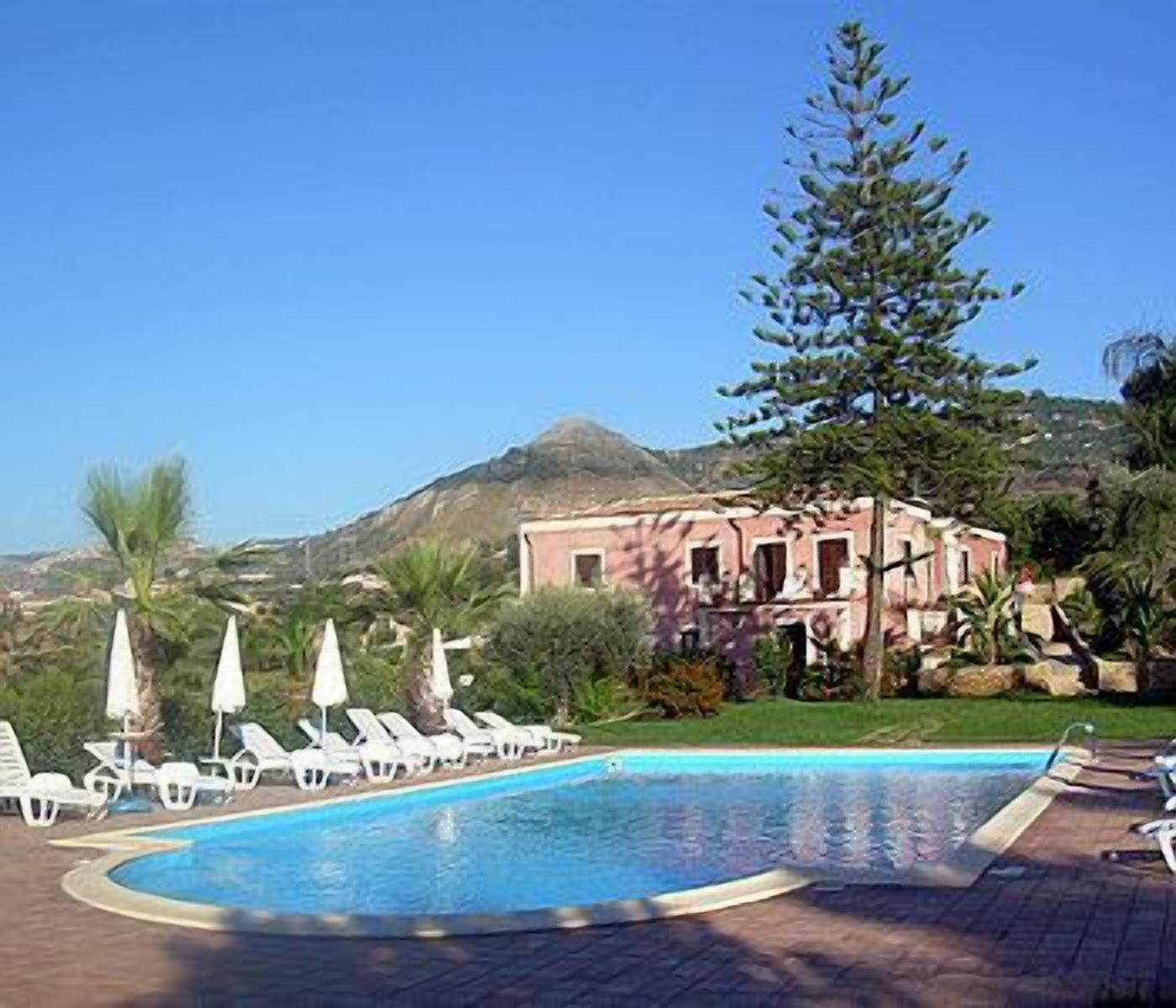 A swimming pool with lounge chairs and umbrellas, a building with a pink facade, and a mountain in the background.