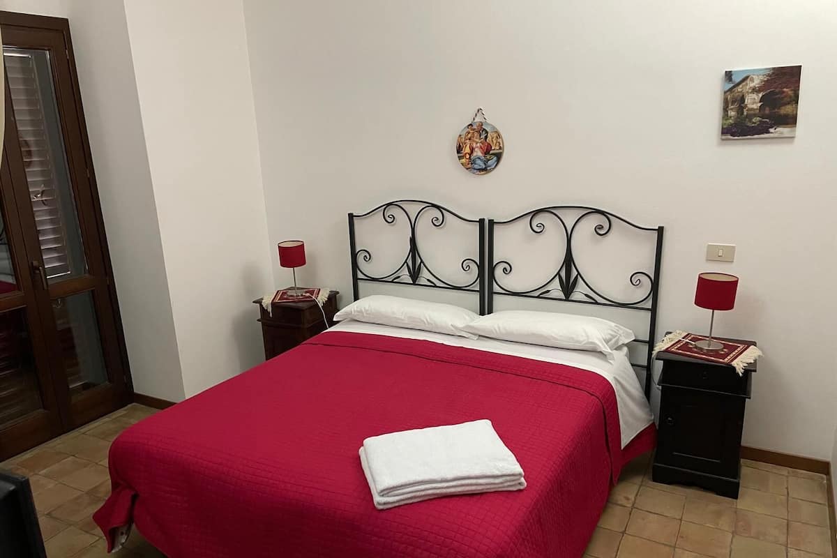 A neatly made bed with a red bedspread and white pillows, a small bedside table with a lamp, and a wall-mounted picture.