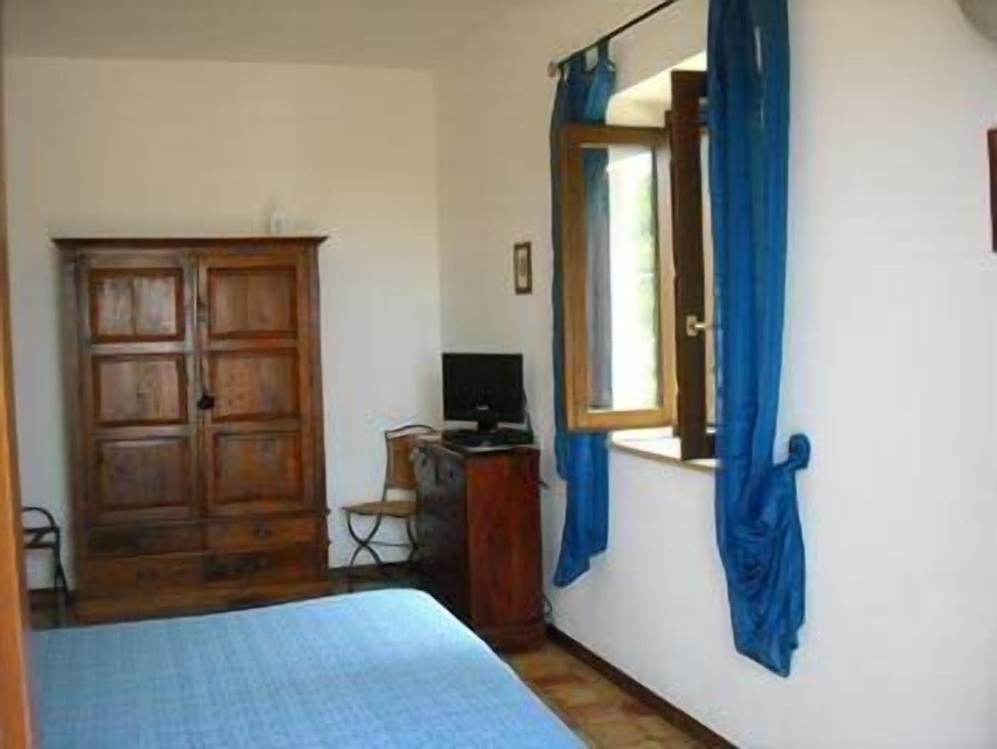 A bedroom with a bed, a wooden wardrobe, a desk with a computer, and a window with blue curtains.