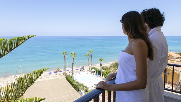 A couple on a balcony overlooking a beach with a pool and palm trees. - Hotel Za' Maria (Caronia)