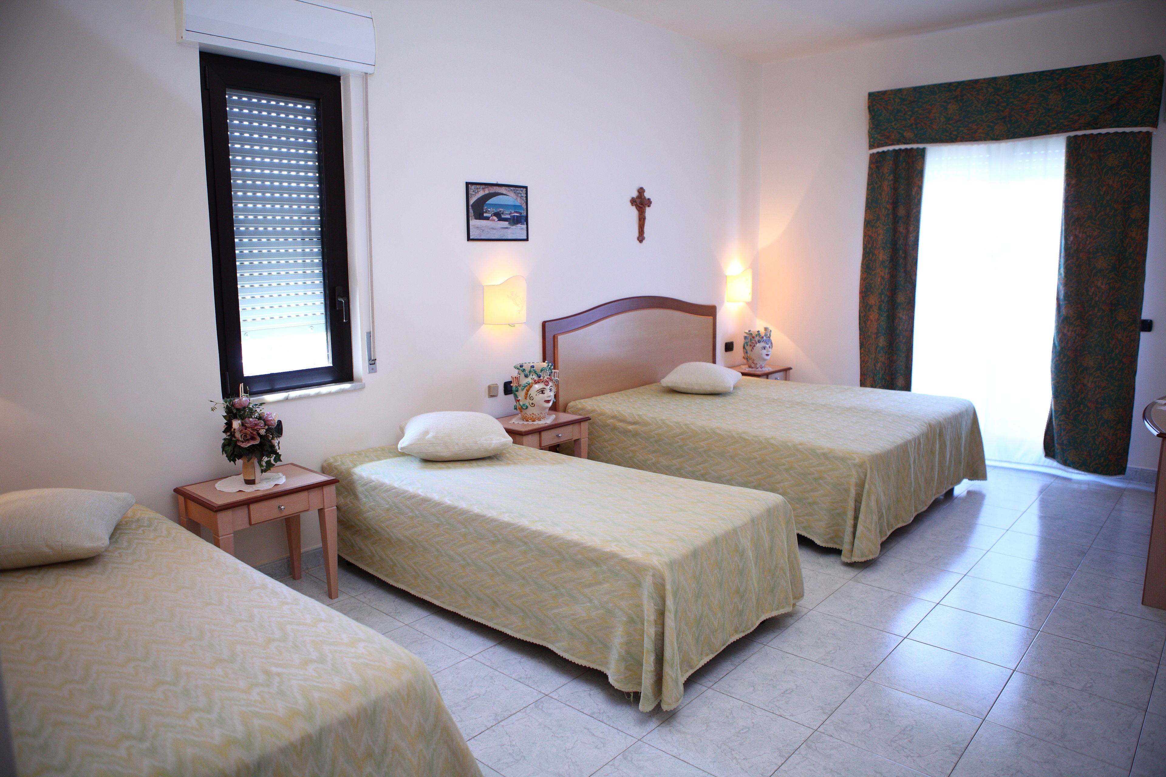 A hotel room with two single beds, a small bedside table with a lamp, a window with blinds, and a picture on the wall.