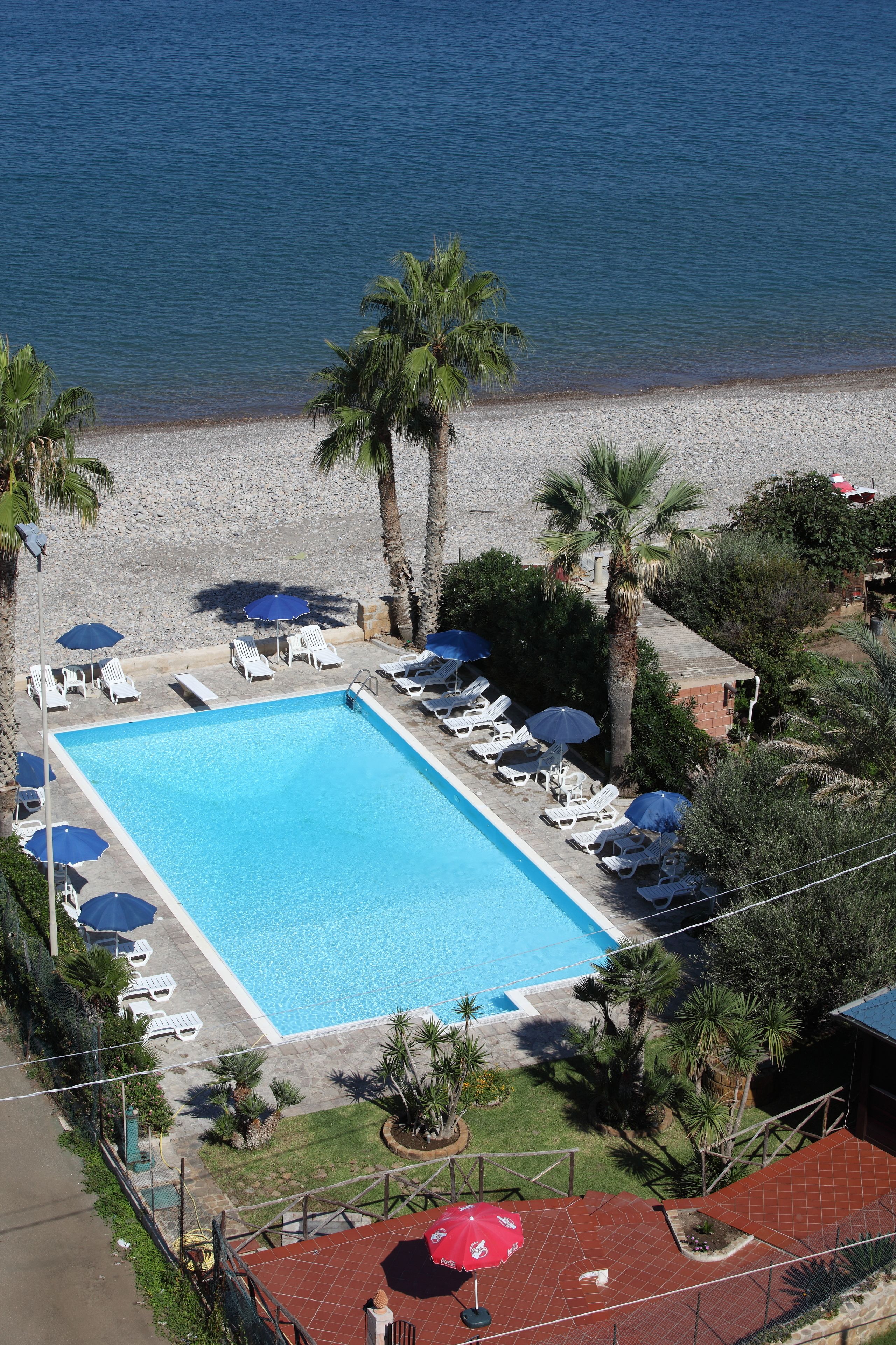 An aerial view of a swimming pool with lounge chairs and umbrellas, surrounded by palm trees and a beach.