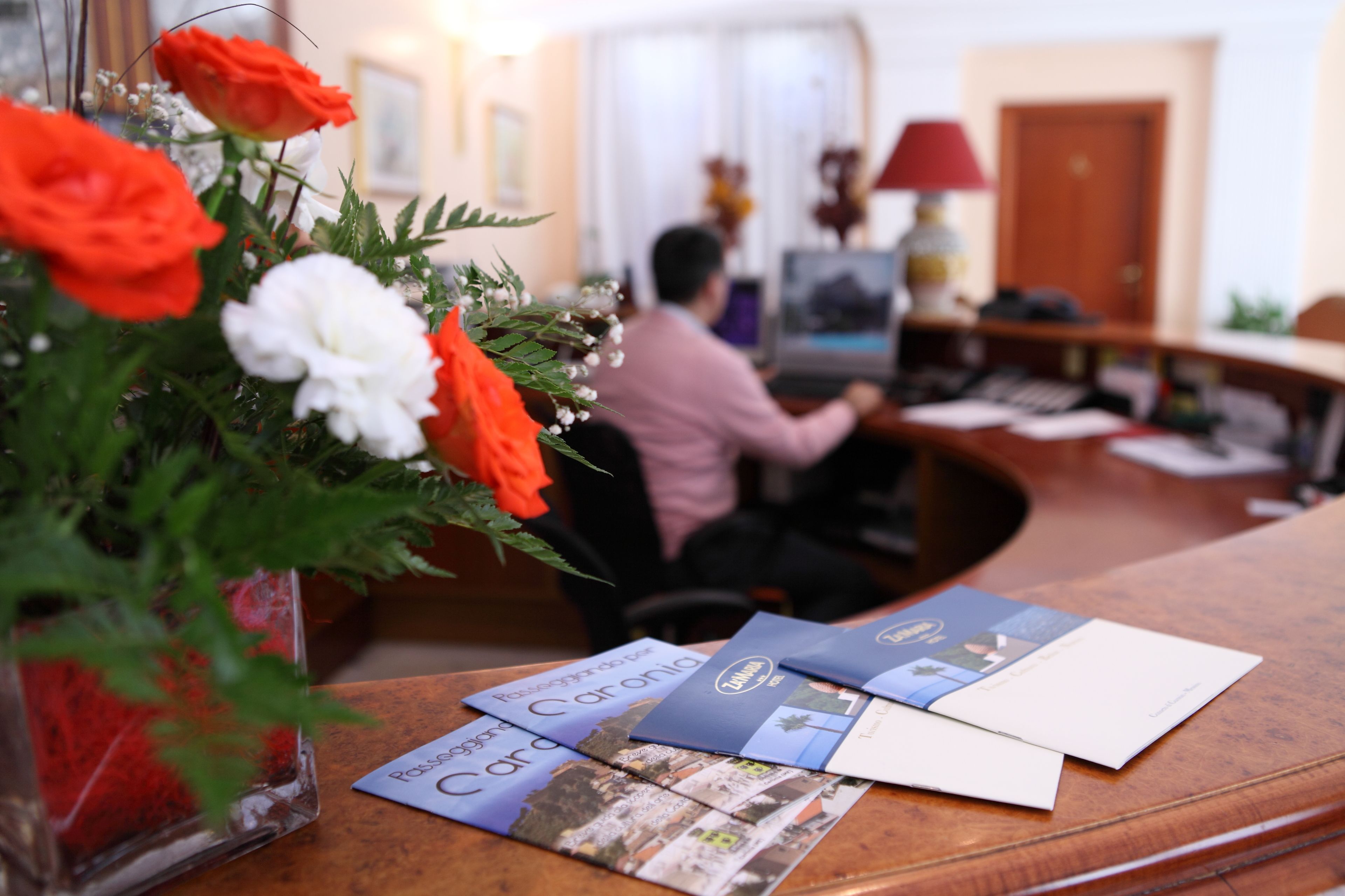 A reception desk with brochures, a vase with flowers, and a person working at the computer.