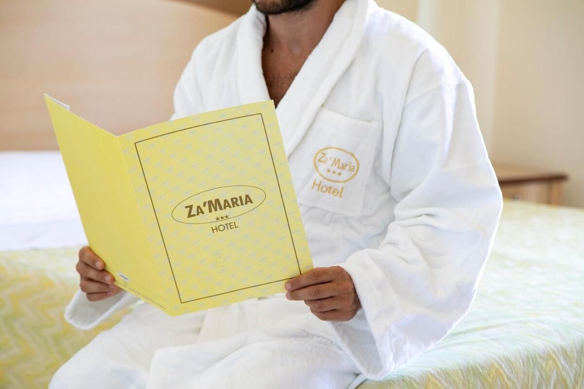 A person in a white robe sitting on a bed with a yellow booklet titled "Za'Maria Hotel".