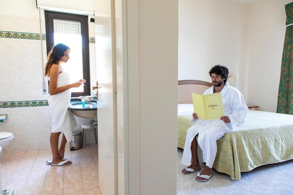A bathroom with a sink, mirror, and toilet. There is a bed with a green bedspread and a person sitting on it reading a book.