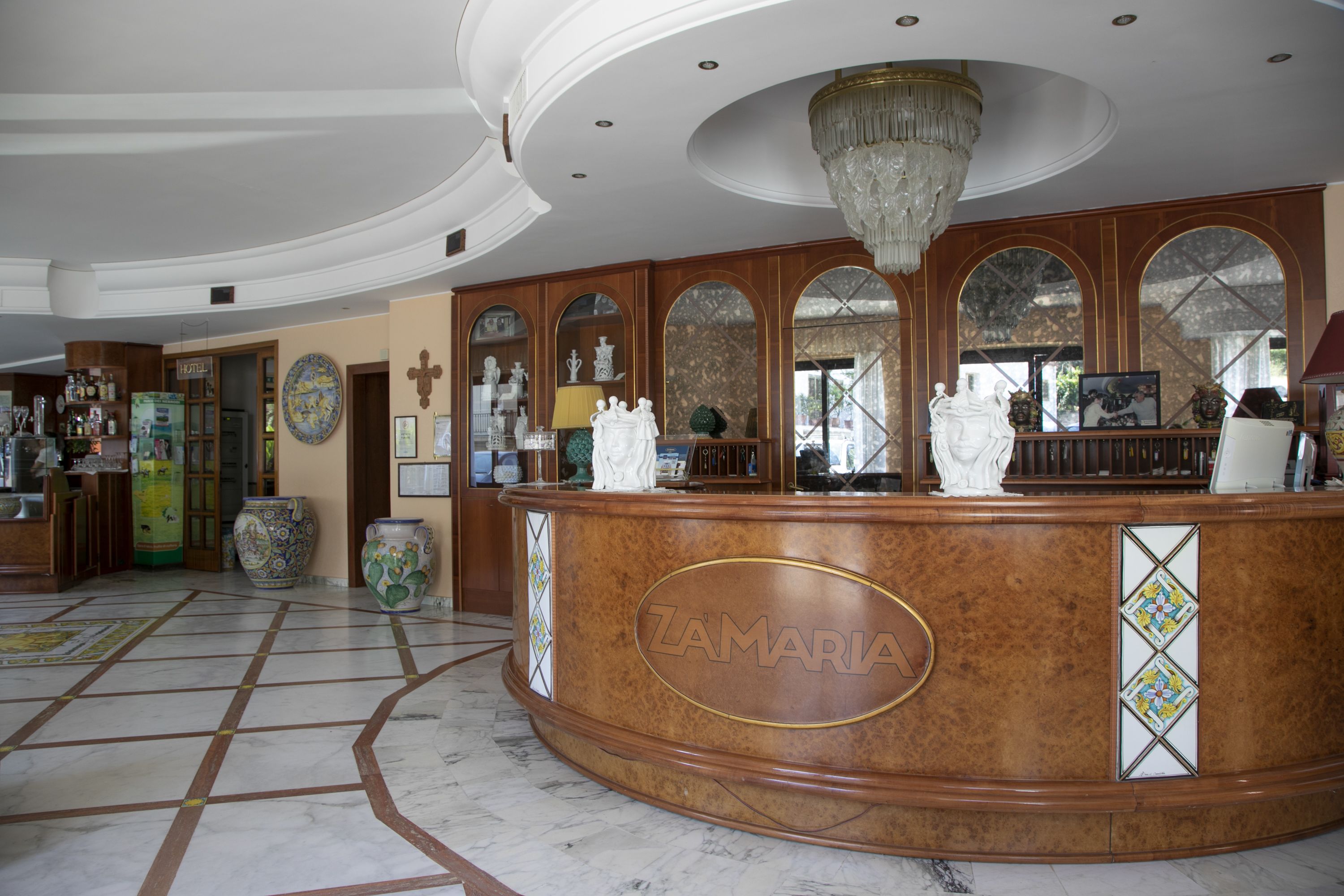 A reception area with a marble floor, a wooden reception desk with the name 'ZAMARIA', and a chandelier.