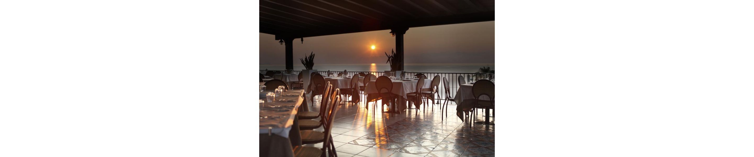 An outdoor dining area with tables and chairs, overlooking a body of water during sunset.