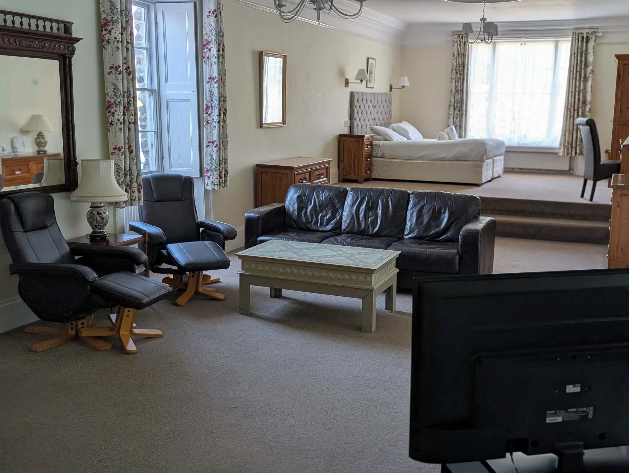A living room with a sofa, chairs, a coffee table, a television, a bed, and a window with curtains.