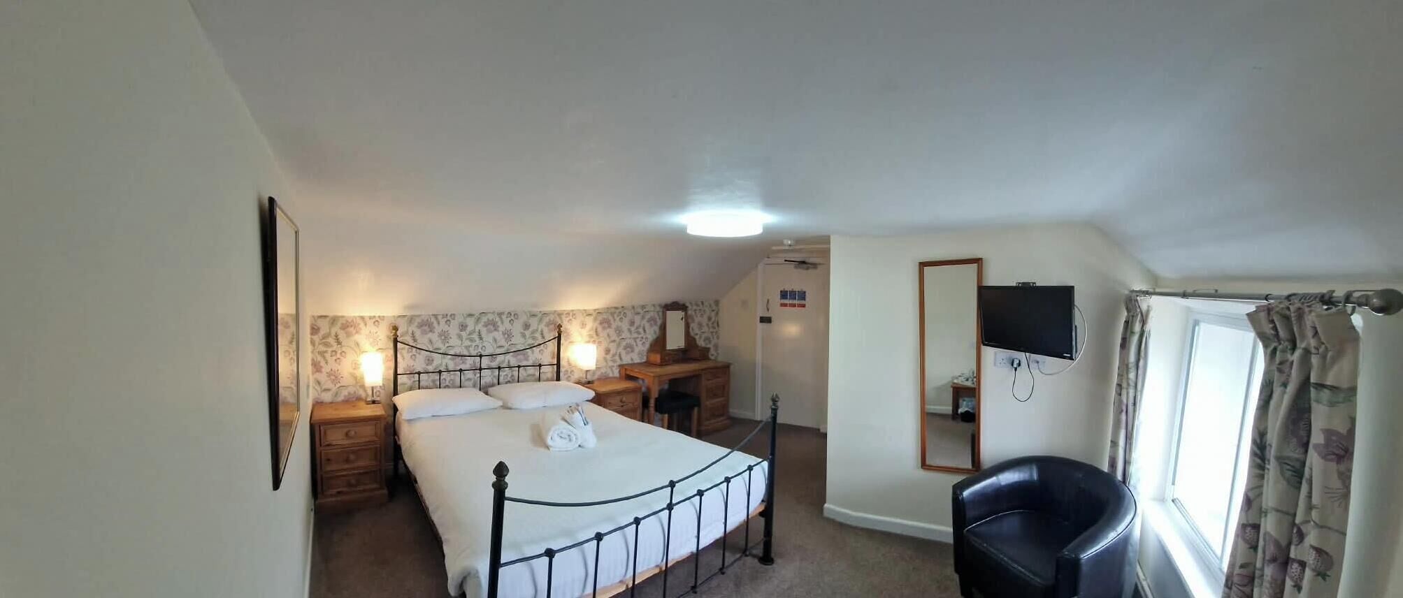 Een slaapkamer met een bed, nachtkastjes, een spiegel, een televisie en een raam met gordijnen.