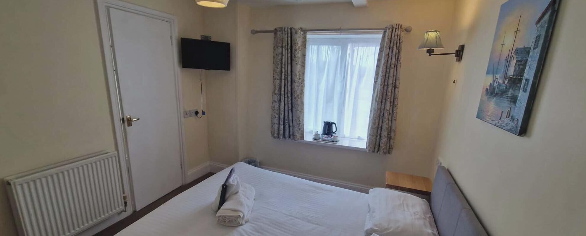 Ett hotellrum med en säng, en tv som hänger på väggen, ett fönster med gardiner, en tavla på väggen och ett element.