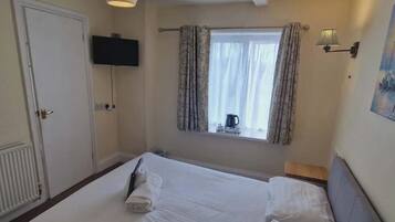 Ett hotellrum med en säng, en tv som hänger på väggen, ett fönster med gardiner, en tavla på väggen och ett element.