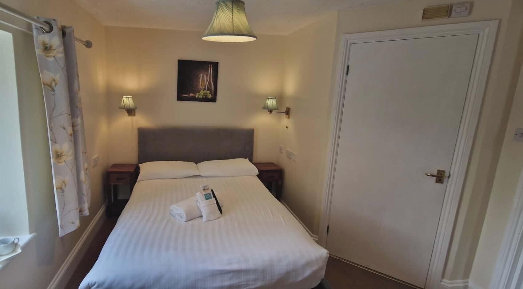 A hotel room with a bed, two bedside lamps, a door, and a window with curtains.