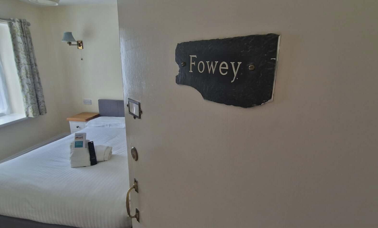A hotel room with a bed, a nightstand, a lamp, and a sign that reads 'Fowey.'