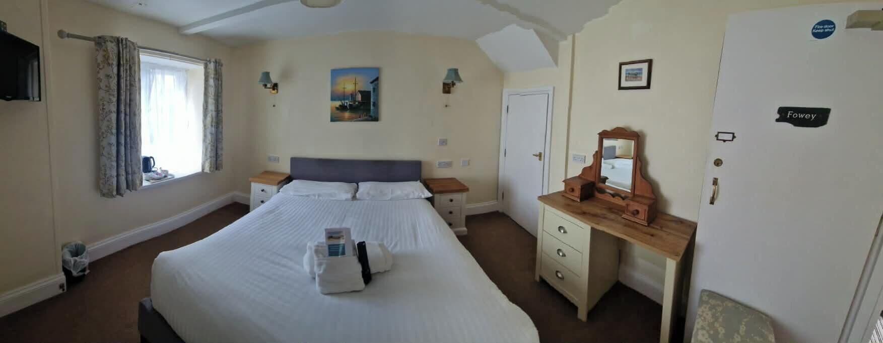 Un dormitorio con cama, mesitas de noche, cómoda, espejo, un cuadro en la pared y una puerta con placa.