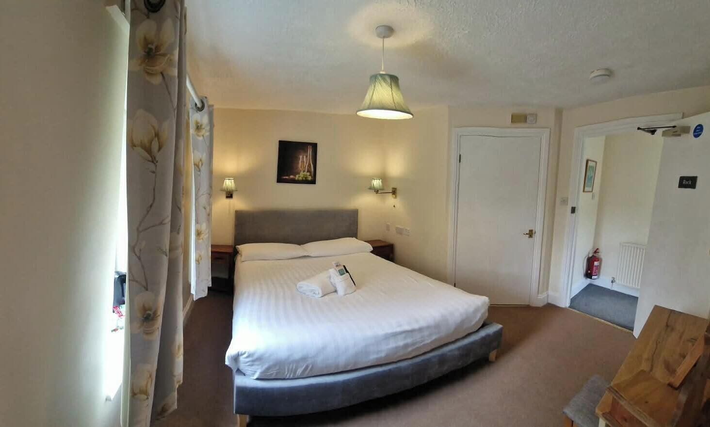 A hotel room with a bed, bedside lamps, a painting on the wall, a door, and a fire extinguisher.