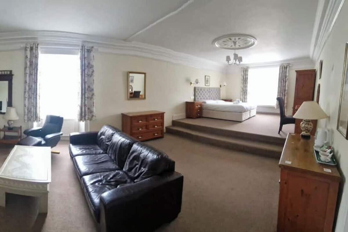 A spacious living room with a black leather sofa, a glass coffee table, a blue armchair, a wooden cabinet, and a bed in the background.