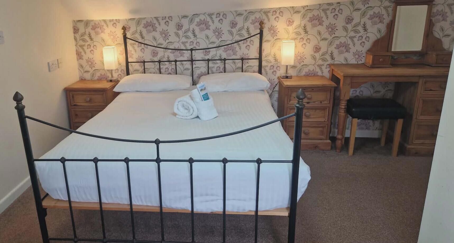 A bed with a metal frame, a wooden nightstand with a lamp, a wooden dresser, and a mirror.