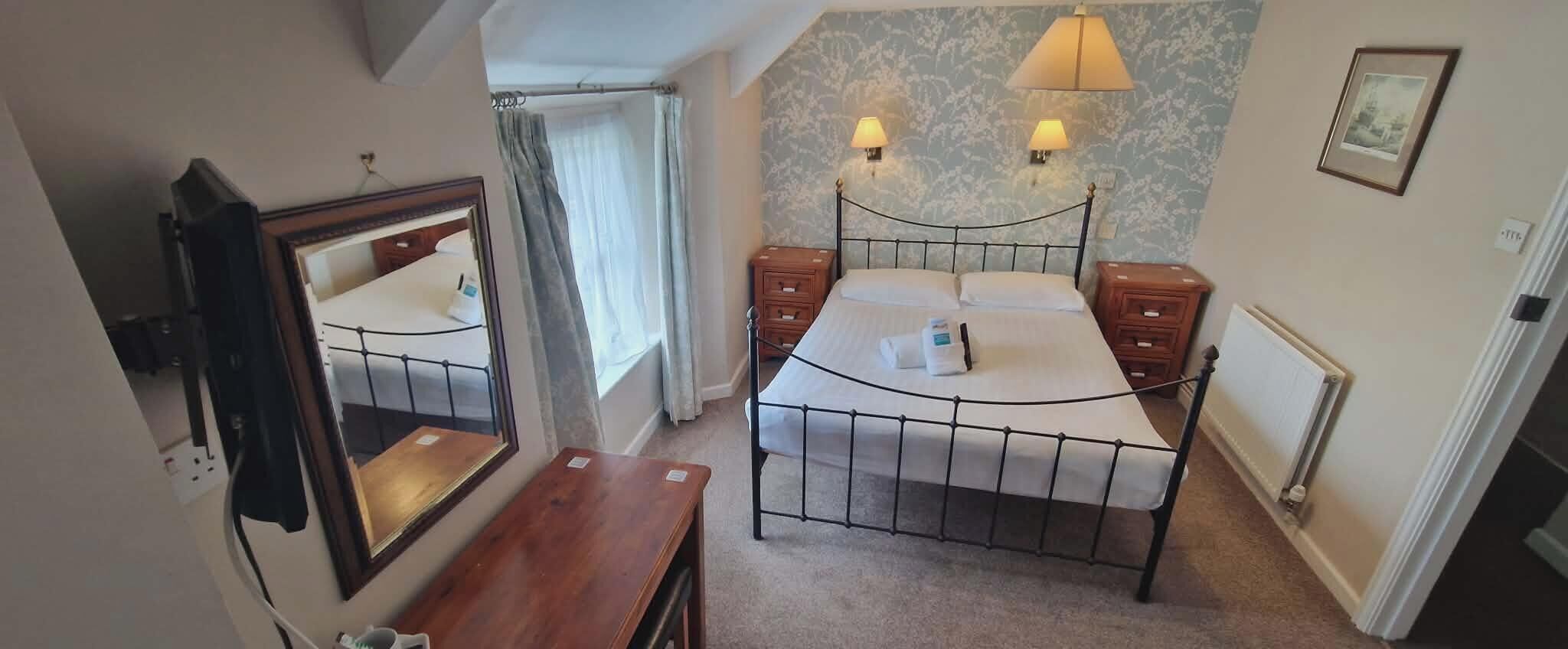 A bedroom with a bed, bedside tables, a mirror, a window with curtains, and a wall with a light fixture.