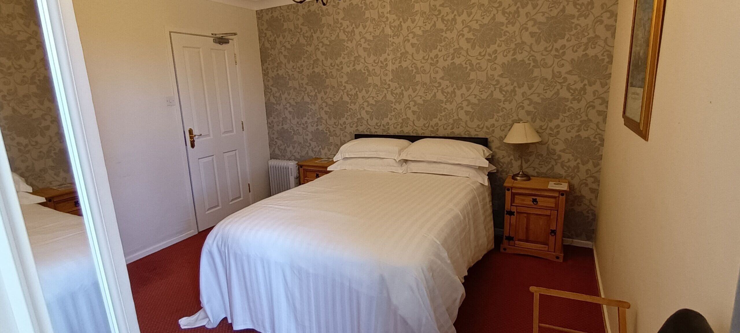 A neatly made bed with white linens, a wooden nightstand with a lamp, and a door with a radiator beside it.
