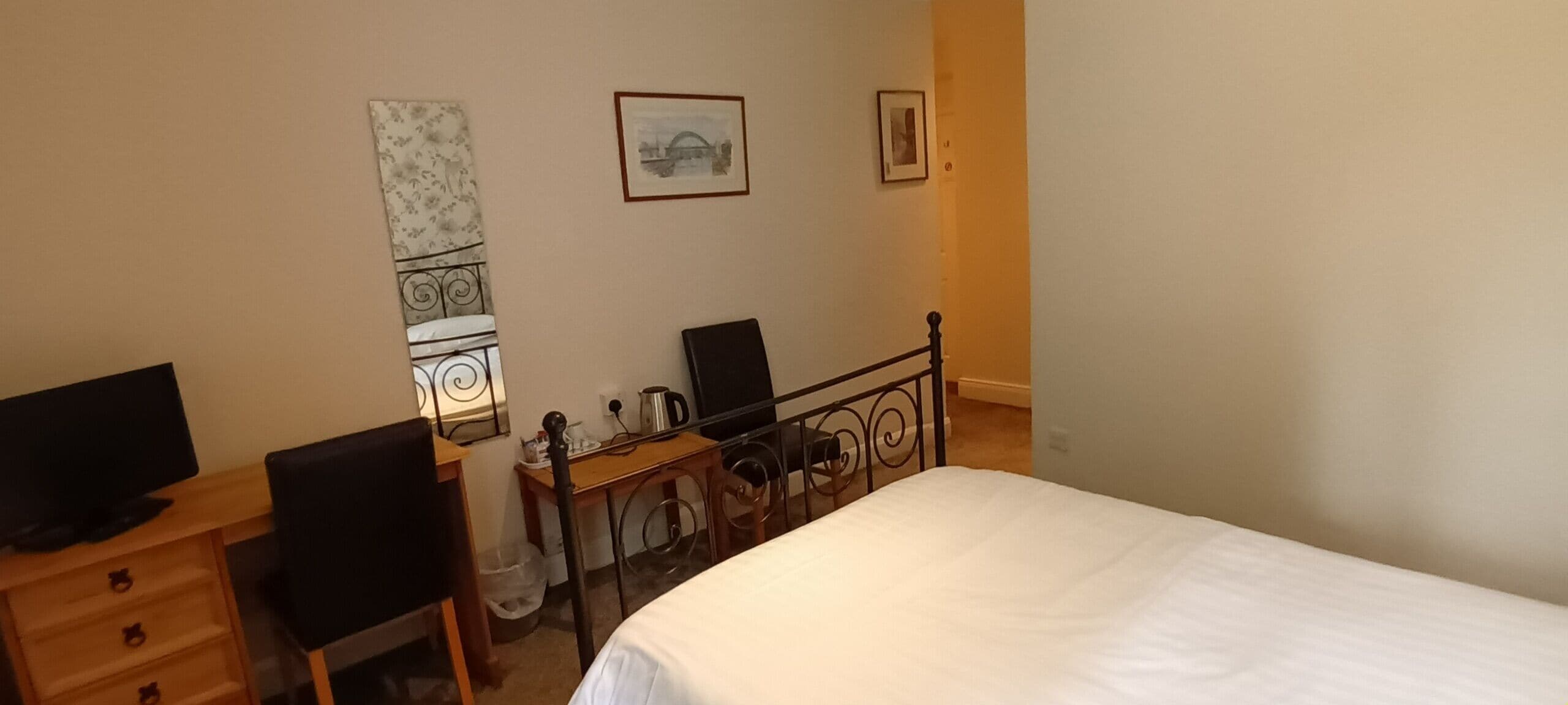 A bedroom with a bed, a desk with a computer, a chair, and a small table with a kettle and cups.