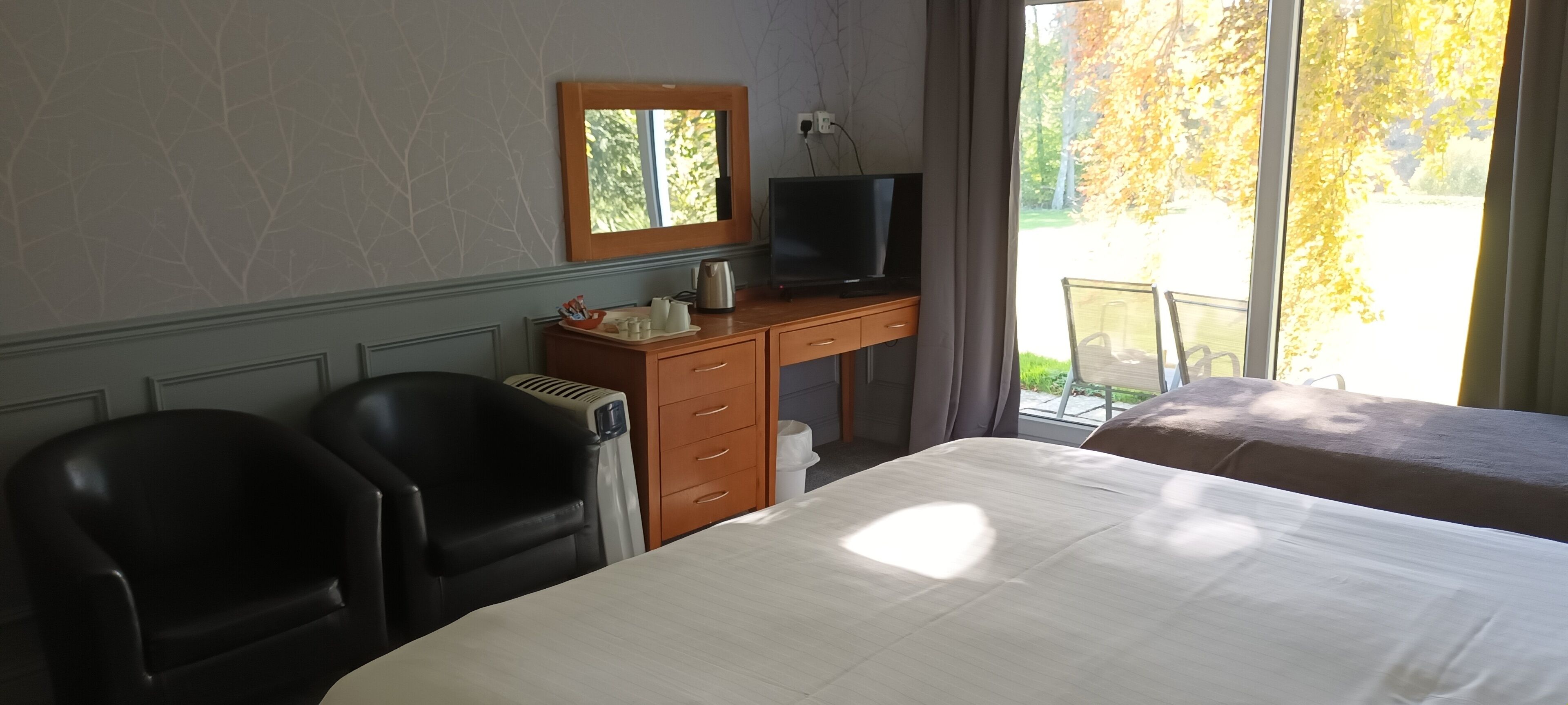 Hotellihuone, jossa on sänky, kaksi tuolia, kirjoituspöytä, jossa on televisio, peili ja ikkuna, josta avautuu näkymä puita kohti.