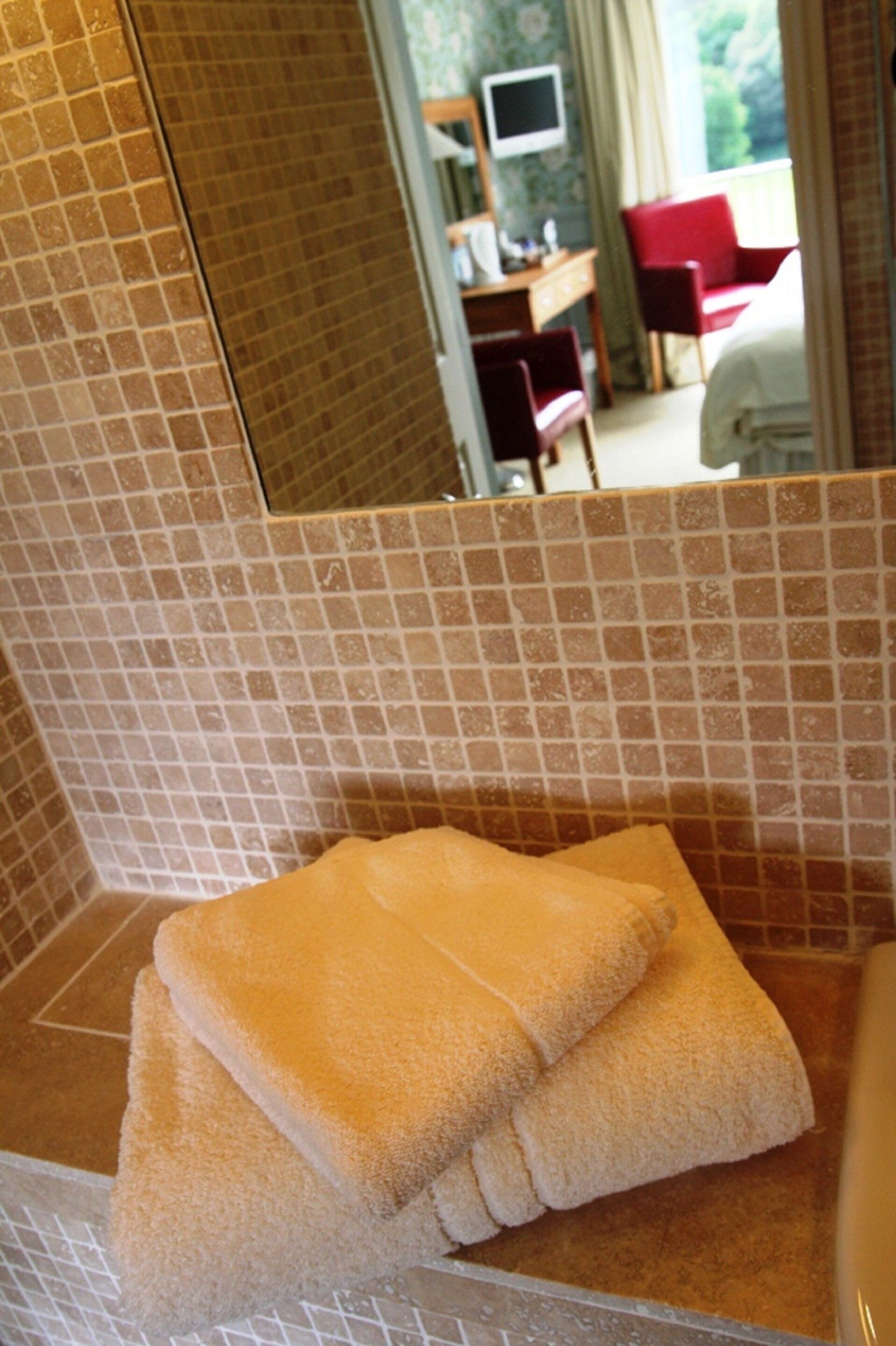 Un baño con pared de azulejos, un lavamanos y un espejo que refleja una habitación con una cama, un escritorio y una silla.