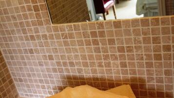 A bathroom with a tiled wall, a sink, and a mirror reflecting a room with a bed, a desk, and a chair.