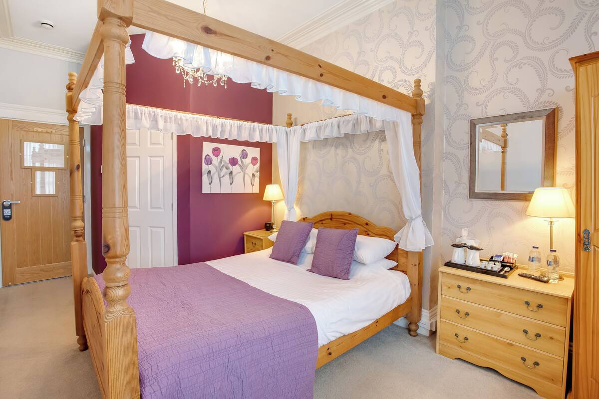 A four-poster canopy bed with a purple bedspread, a wooden nightstand with a lamp, and a dresser with drawers.