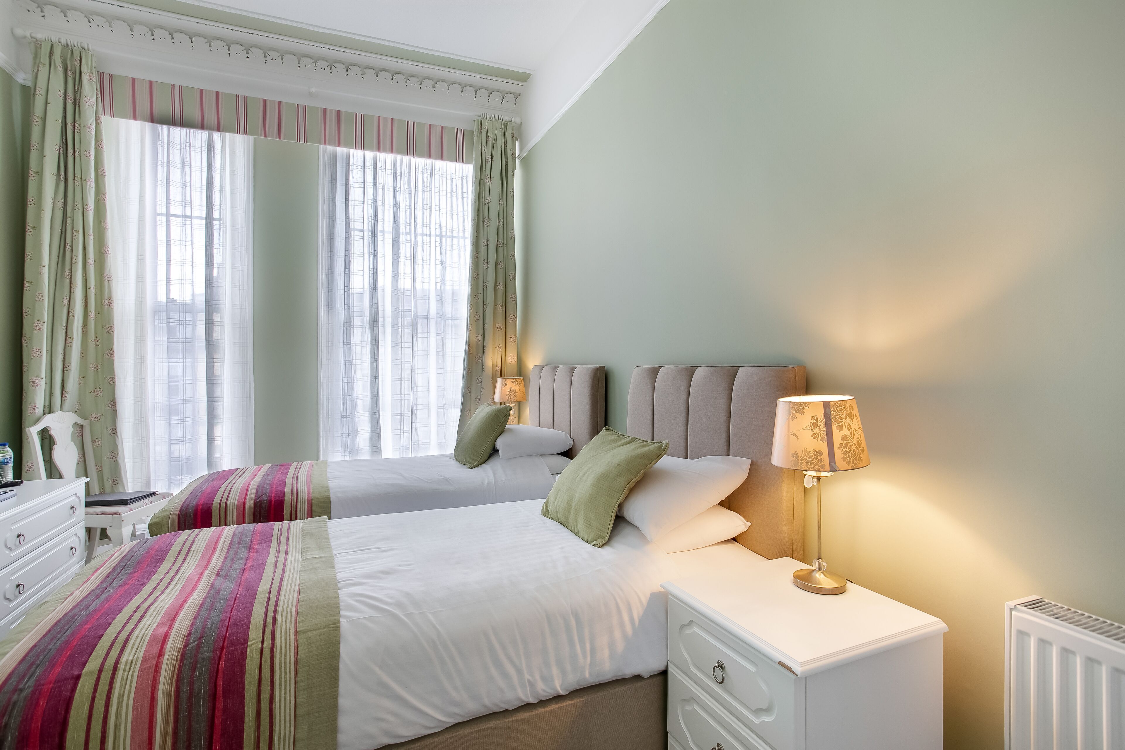Una habitación de hotel con dos camas, una mesita de noche con lámpara, un aparador y una ventana con cortinas.
