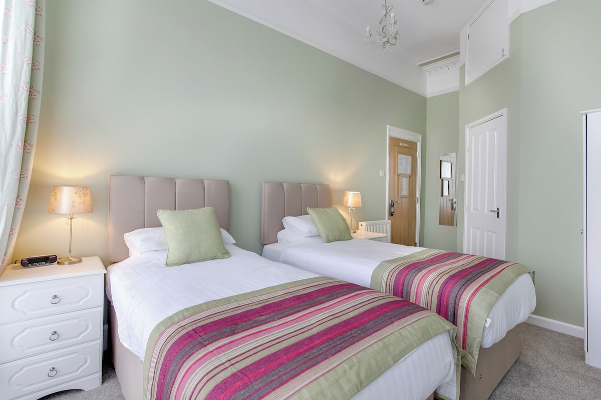 A hotel room with two beds, a nightstand, a lamp, and a door.