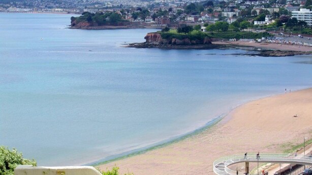 A coastal view with a sandy beach, a rocky outcrop, and a cityscape in the background.