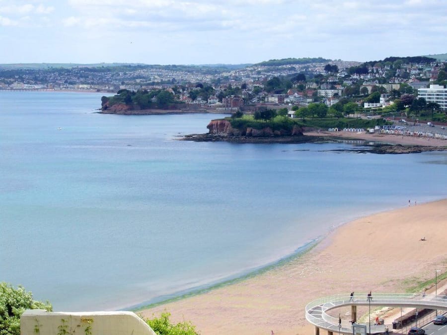 Vista costeira com praia de areia, um afloramento rochoso e paisagem urbana ao fundo.