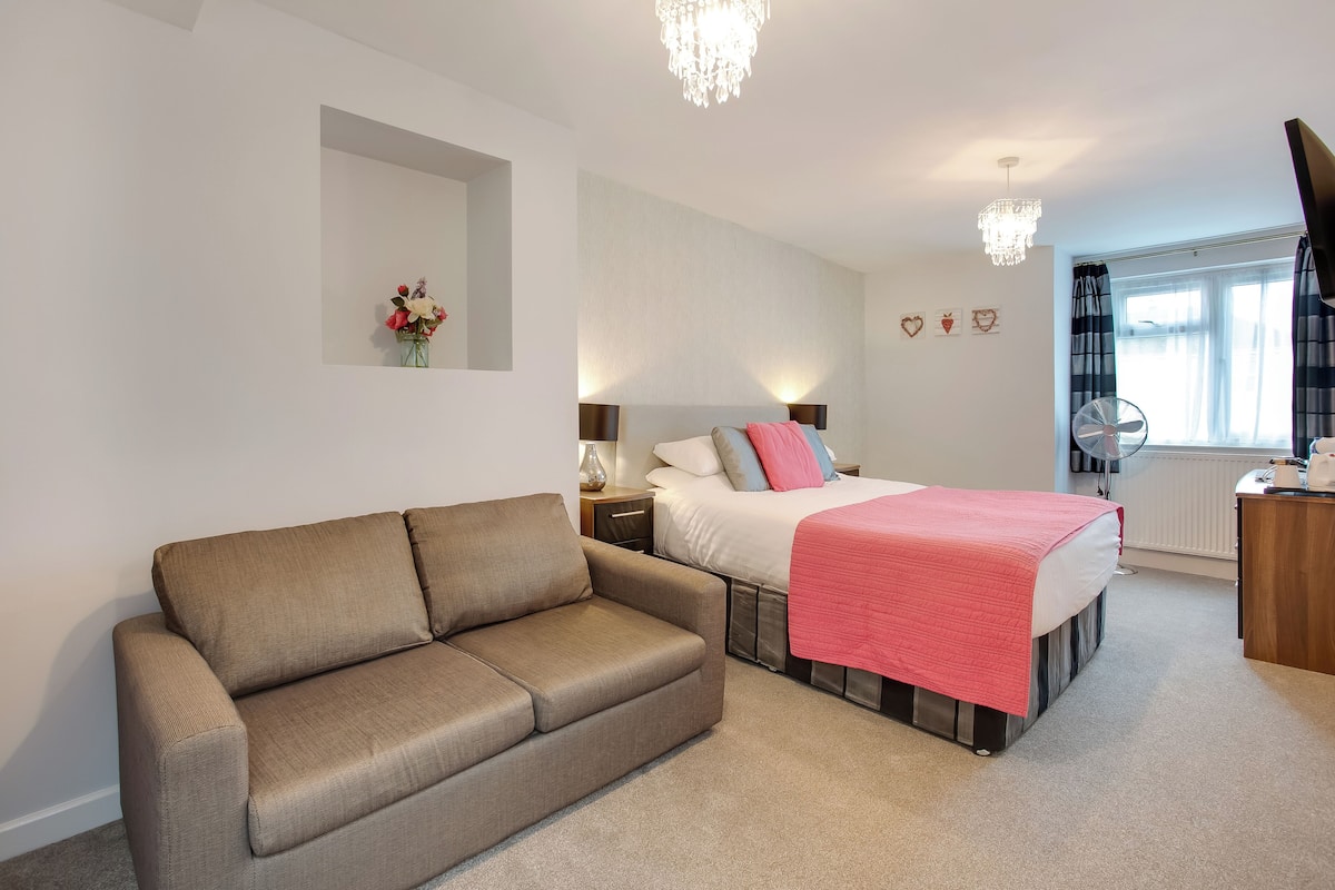 A bedroom with a bed, a sofa, a bedside table, a mirror, a chandelier, a window with curtains, and a radiator.