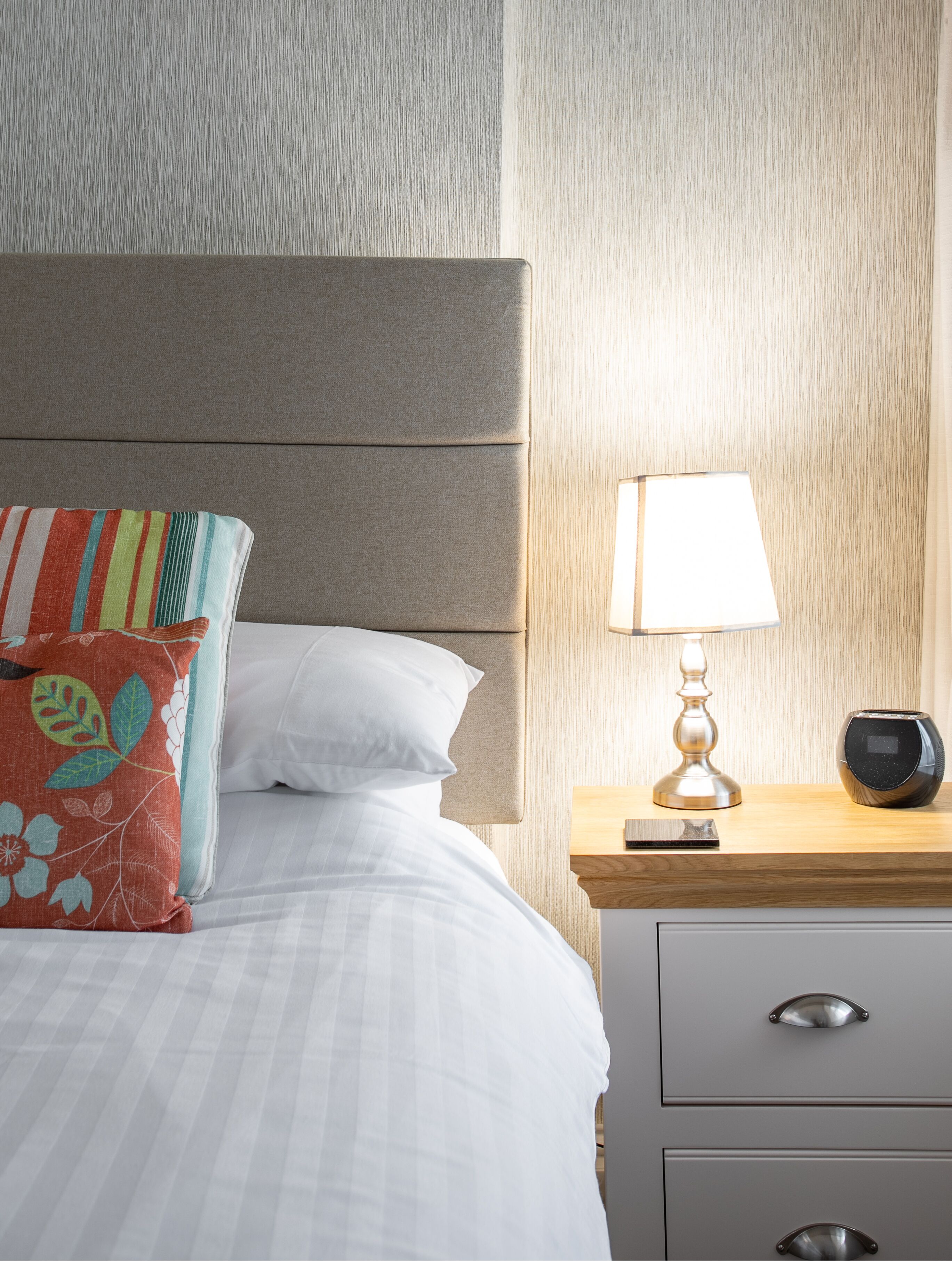 A neatly made bed with a striped pillow, a bedside lamp, and a speaker on a wooden nightstand.