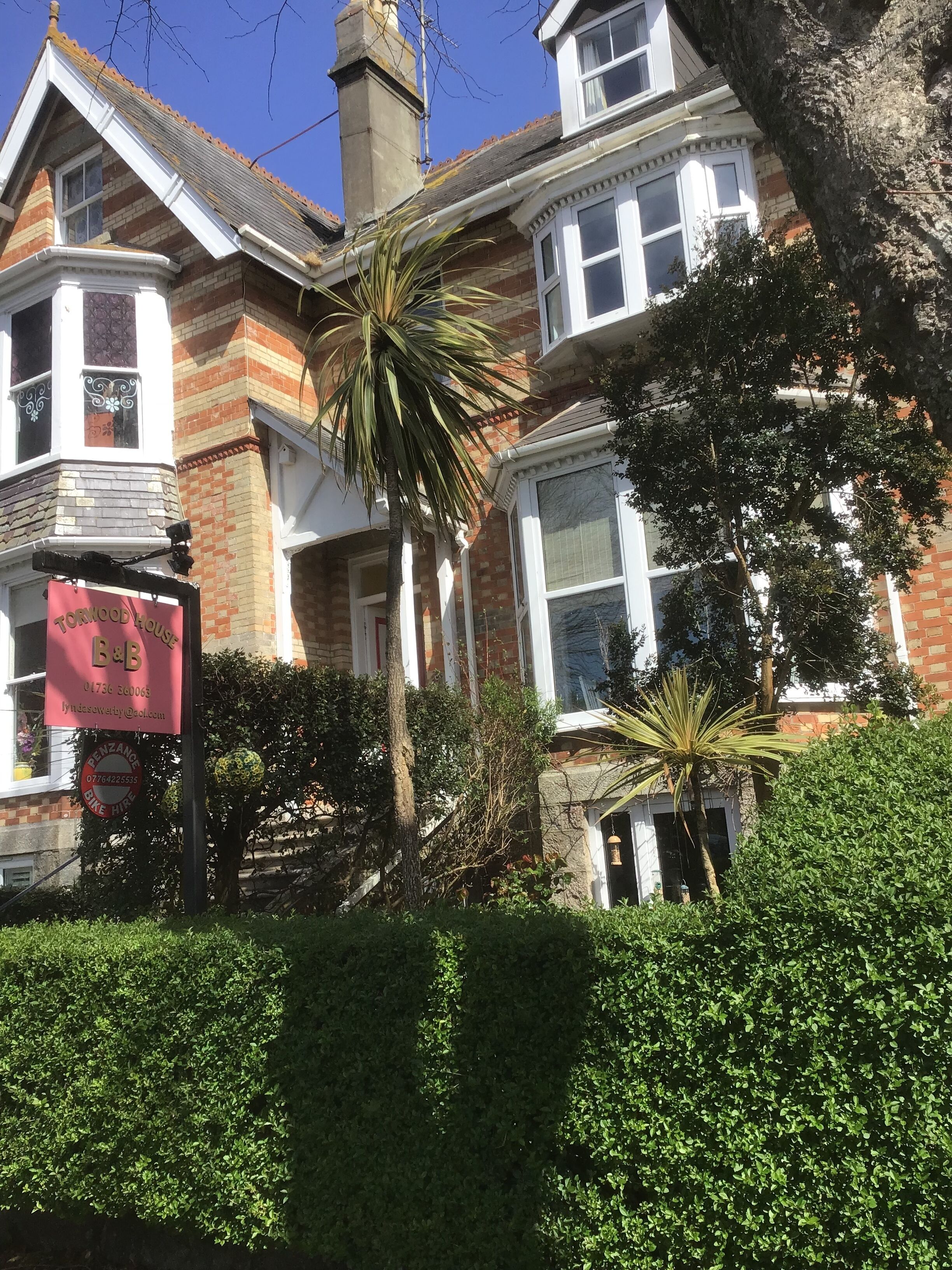 A traditional brick house with a bay window, a red sign for a B&B, and a well-maintained garden with various plants and a palm tree.