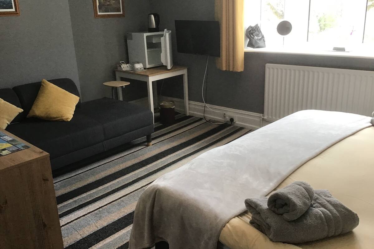A hotel room with a bed, a sofa, a small table, a radiator, and a window with curtains.