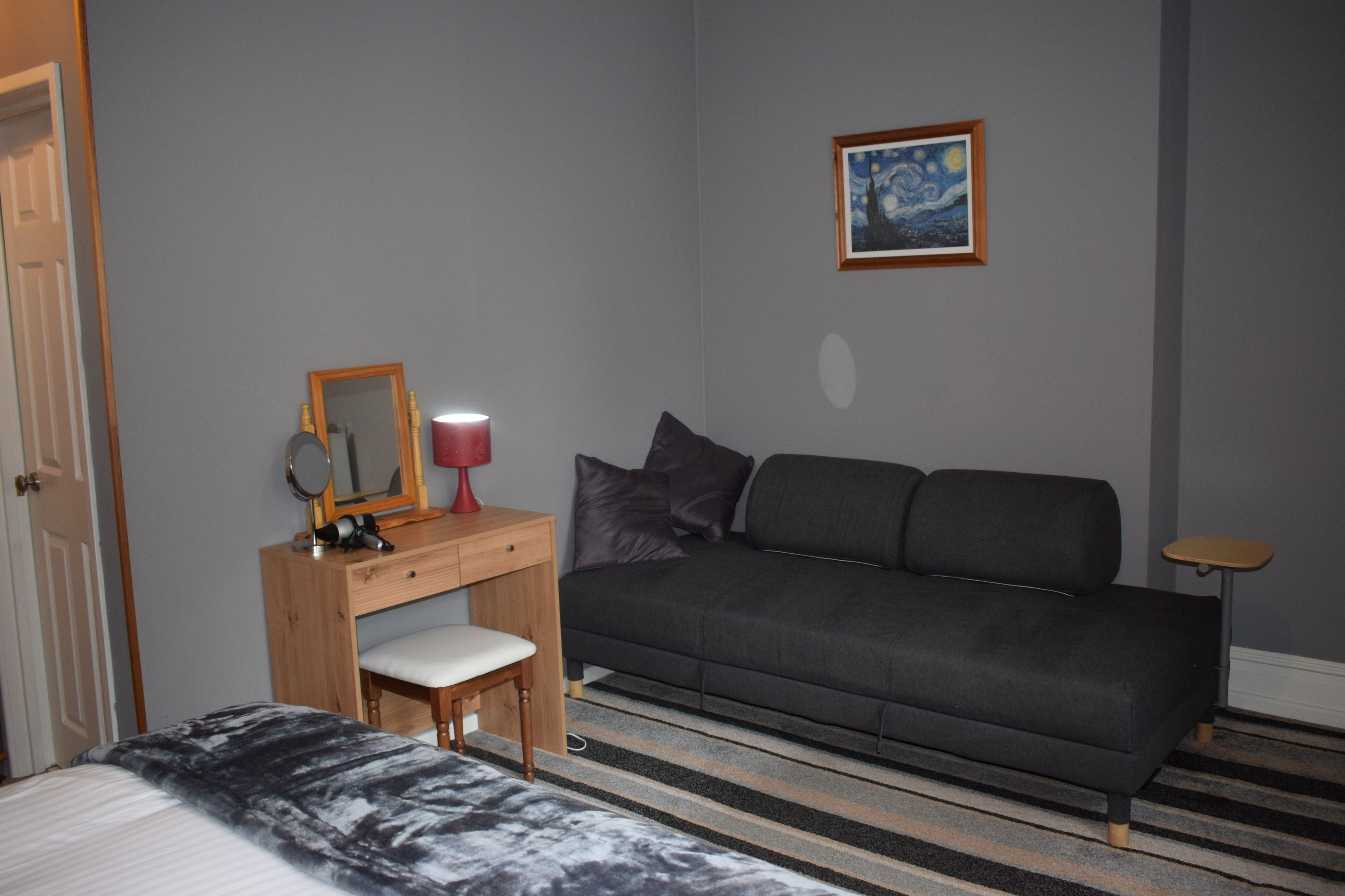 Un dormitorio con cama, escritorio con silla, sofá gris, una mesita, lámpara, espejo y un cuadro en la pared.