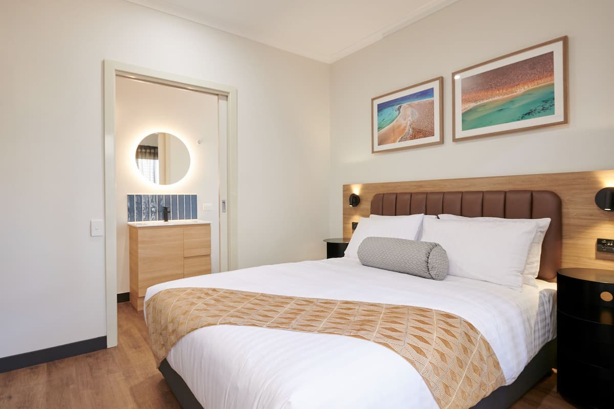 A neatly arranged bedroom with a large bed, a bedside table, a nightstand, a mirror, and two framed pictures on the wall.