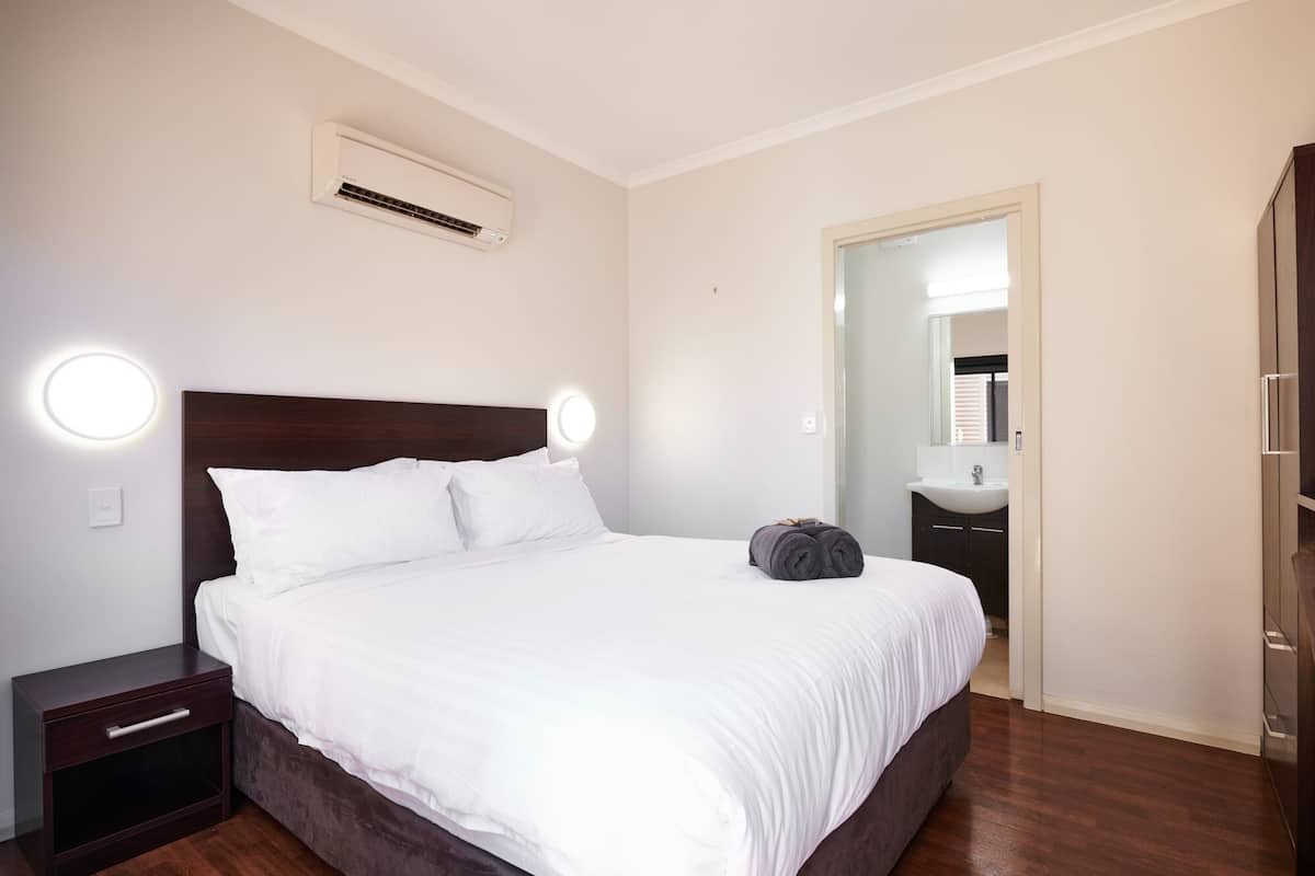 A bedroom with a bed, bedside tables, an air conditioner, and a bathroom visible through an open door.