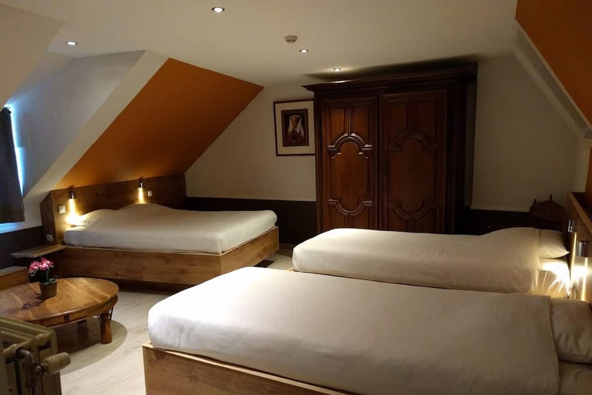 Quarto de hotel com duas camas, um guarda-roupa de madeira, uma mesa redonda pequena com um vaso de flores e um quadro na parede.