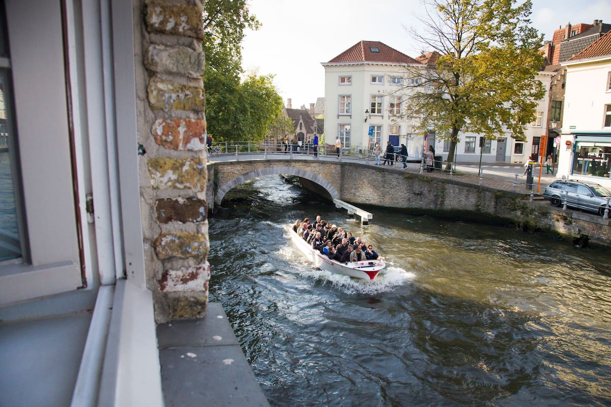 Um canal estreito com um barco transportando passageiros, uma ponte de pedra e edifícios ao longo da margem.
