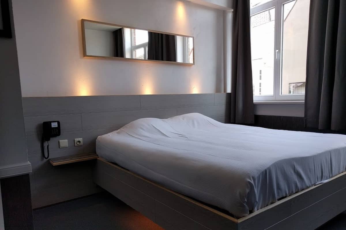 Quarto de hotel moderno com uma cama grande, telefone ao lado da cama, espelho fixado na parede e janela com cortinas.