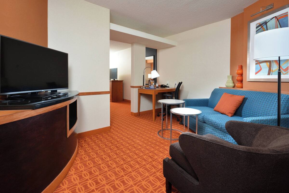 A hotel room with a TV, a desk with a lamp, a blue patterned sofa, and a brown armchair.