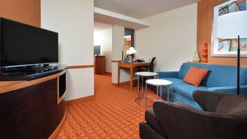 A hotel room with a TV, a desk with a lamp, a blue patterned sofa, and a brown armchair.